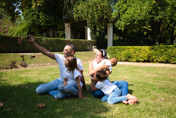 rodzinny portret pięciu członków, ojca, matki i dzieci siedzących na trawie, robiących zdjęcia telefonem komórkowym, podczas gdy jesienią liście spadają z drzew. Koncepcja rodziny. - Zdjęcie, obraz