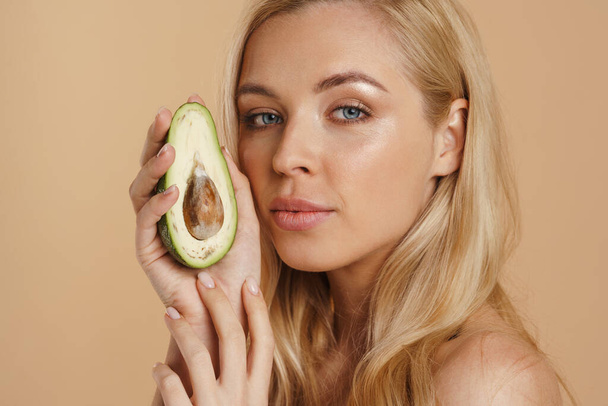 Smiling young blonde topless woman with long hair standing over beige background, holding avocado  - Valokuva, kuva