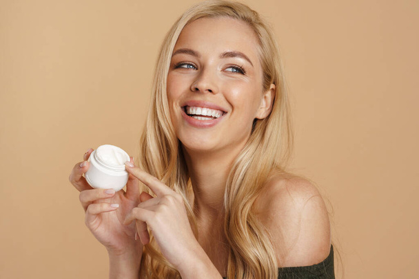 Smiling young blonde woman with long hair showing facial cream jar standing over beige background - Fotografie, Obrázek