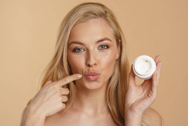 Smiling young blonde white woman beauty portrait isolated over beige background holding jar of facial cream - Φωτογραφία, εικόνα