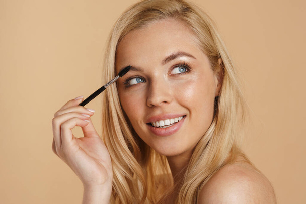 Smiling young white blonde woman with bare shoulders standing over beige background, holding eyebrow brush - Fotografie, Obrázek