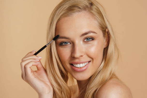 Smiling young white blonde woman with bare shoulders standing over beige background, holding eyebrow brush - Foto, imagen