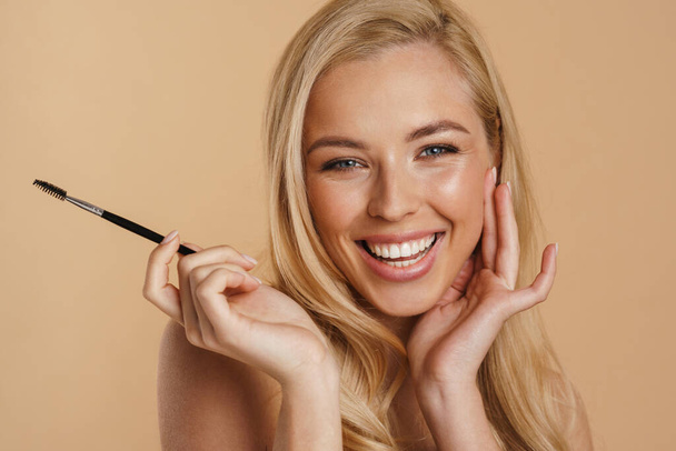 Smiling young white blonde woman with bare shoulders standing over beige background, holding eyebrow brush - Φωτογραφία, εικόνα