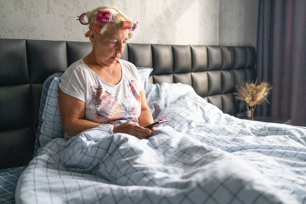 Femme caucasienne adulte utilisant le téléphone mobile vérifier le flux de réseau social ou chat message assis dans le lit à la maison. - Photo, image