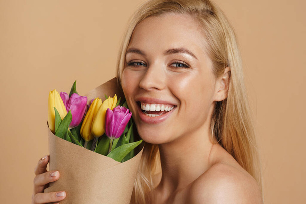 Happy young blonde white woman with long hair standing over beige background with bare shoulders holding bouquet of tulips - Photo, image