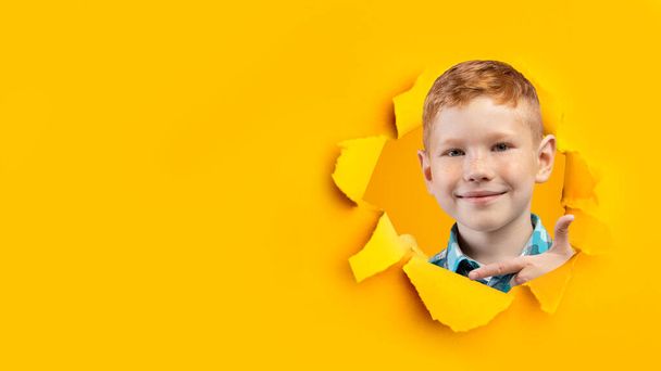Cute red haired little boy looking through hole in ripped orange paper, pointing aside at empty space, banner design - Fotó, kép