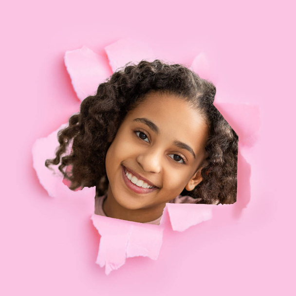 alegre sonriente africano americano niña mirar a través de agujero en rosa papel - Foto, Imagen