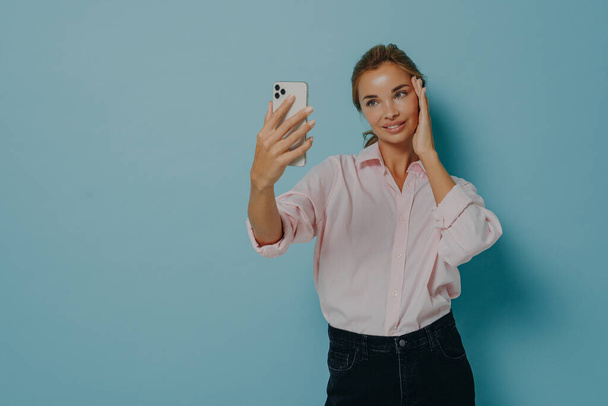 Элегантная юная леди в повседневной одежде пытается сделать селфи, держа в руках смартфон, изолированный на голубом - Фото, изображение