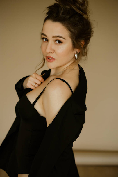 portrait of young beautiful caucasian woman in a black jacket , slender beautiful lady with hairstyle and makeup in a sexy image, stylish serious brunette in a business suit on beige background. - 写真・画像