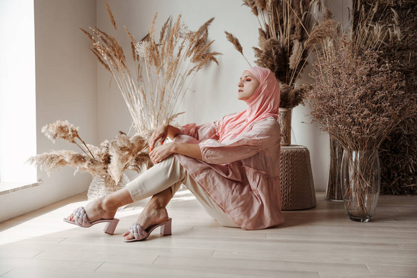Hermosa mujer árabe musulmana usando hijab rosa sentada en el suelo y sonriendo - Foto, Imagen