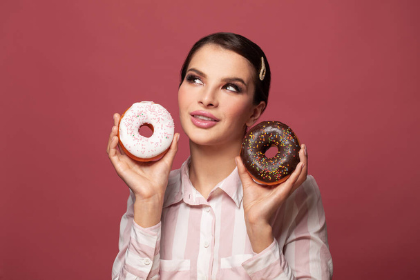 Sweet snack, delicious pastry. Cute smiling woman with fancy dark hair looking up and holding donut, having fun with doughnut, showing tasty dessert on red background - Фото, изображение
