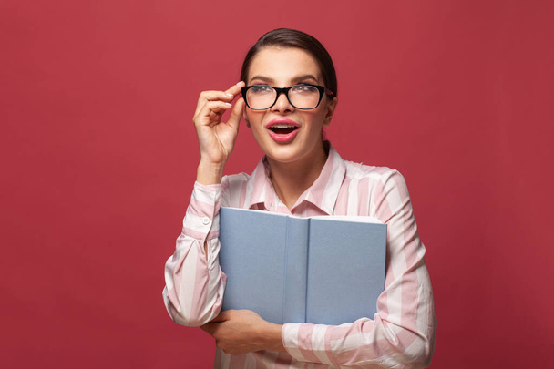 Portrait young happy surprised woman in glasses holds book. Reading, education, studying and learning knowledge concept. Student smiling, positive emotion on red background - 写真・画像