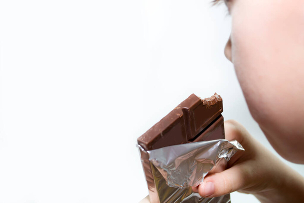 The boy holds a bar of chocolate and eats on a white background. - Fotografie, Obrázek
