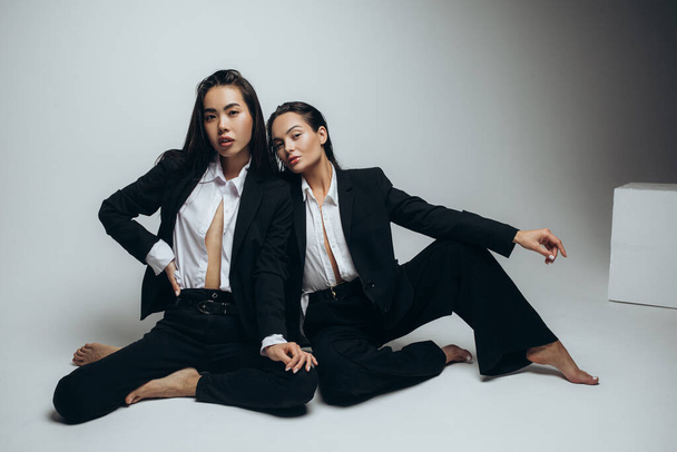 Zwei junge brünette Frauen posieren im Studio sitzend in schwarzen Anzügen. - Foto, Bild