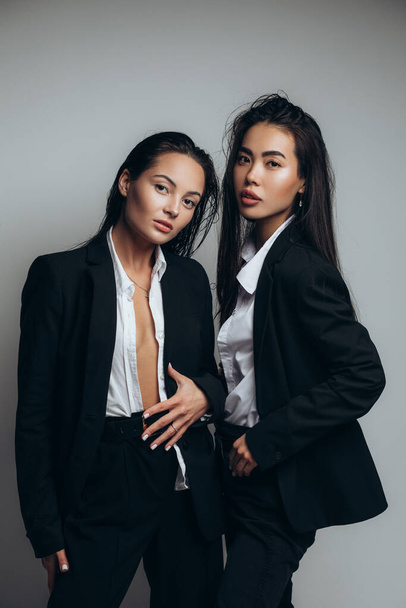 Δύο νεαρές μελαχρινές γυναίκες ποζάρουν σε στούντιο ντυμένες με μαύρα κοστούμια. - Φωτογραφία, εικόνα