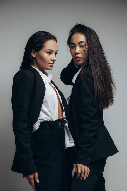 Two young brunette women pose in studio standing dressed in black suits. - Foto, Imagen
