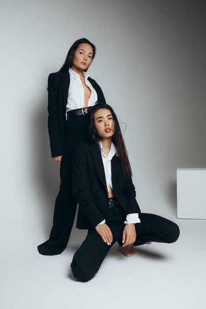 Two young brunette women pose in studio standing dressed in black suits. - Photo, Image