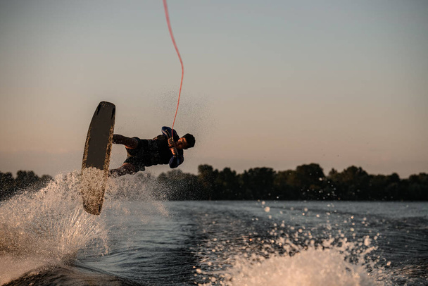 male wakeboarder making trick in jump with wakeboard over splashing river. Summertime watersports activity - Photo, image