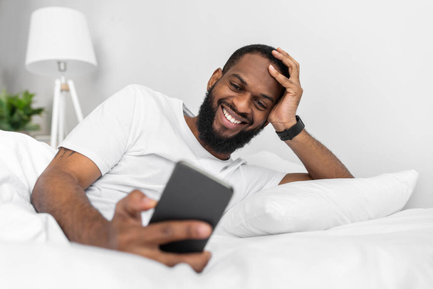 Χαρούμενος νεαρός Αφροαμερικάνος με γενειάδα κοιτάζει το smartphone, ξαπλώνει στο λευκό κρεβάτι. - Φωτογραφία, εικόνα