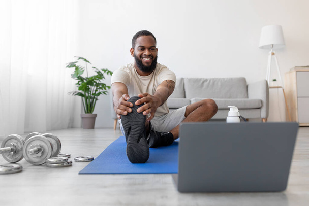 Cheerful young african american guy with beard doing stretching for legs on mat and look at laptop - Fotó, kép