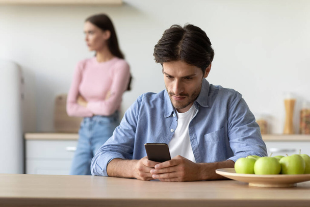Busy millennial european guy plays in game in smartphone and ignores offended wife at kitchen - Fotó, kép