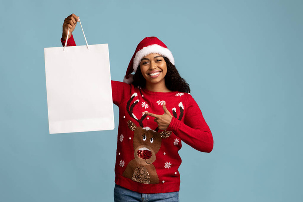 Cheerful lady in Santa hat pointing at white shopping bag - Zdjęcie, obraz