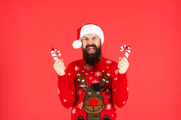 shouting bearded man in santa claus hat and sweater hold candy stick on red background, new year - Foto, afbeelding