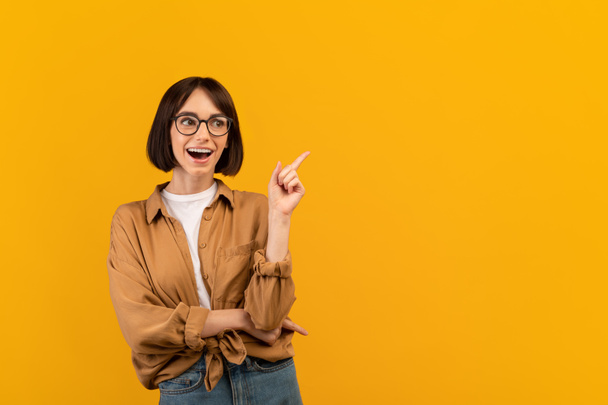Overjoyed young woman pointing finger aside advertising text standing over yellow studio background, empty space - Photo, Image