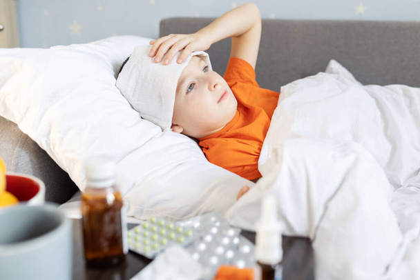 Ein kranker Junge liegt mit Fieber bedeckt im Bett, neben dem Medikament auf dem Tisch. Grippe-Saison. - Foto, Bild