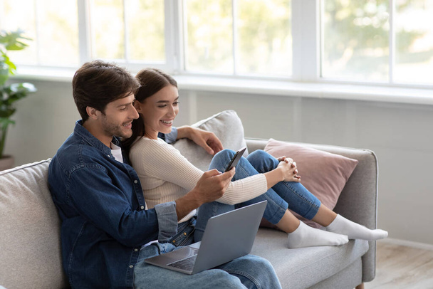Happy millennial european male and female look at smartphone and call online in living room interior - Photo, Image