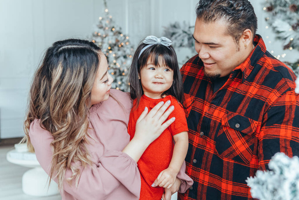 Asian family father, mother with daughter toddler girl celebrating Christmas or New Year. Mixed race mom, dad, daughter decorating Christas tree hugging at home. Winter holiday celebration. - Photo, Image