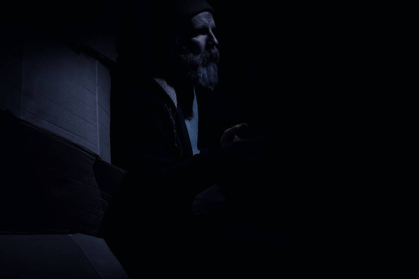 Un hombre sin hogar barbudo se sienta en cajas en la calle y pide ayuda. ¿Necesita una persona sin hogar pide dinero para la comida y durante la noche. - Foto, imagen
