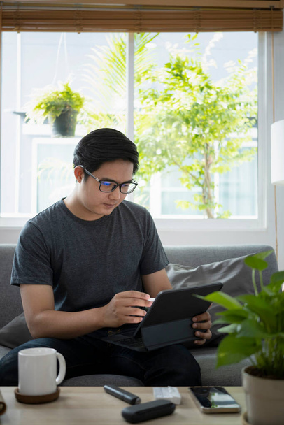 Mladý asijský muž pracující on-line s počítačem tablet zatímco sedí na gauči. - Fotografie, Obrázek