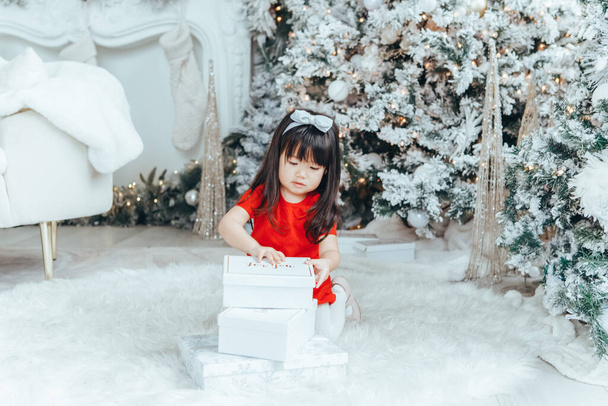 Asian toddler girl in red dress opening gift boxes with presents under Christmas tree at home. Kid child celebrating Christmas or New Year. Winter holiday celebration. - Photo, Image