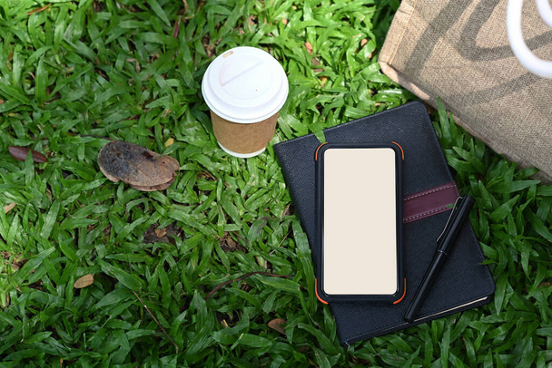 Mock up smart phone, koffiebeker en boek op groen gras in het natuurpark. - Foto, afbeelding