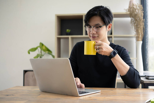 Young asian man drinking hot coffee and checking news on laptop computer. - Foto, Imagen