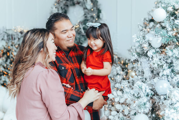 Asian family father, mother with daughter toddler girl celebrating Christmas or New Year. Mixed race mom, dad, daughter by decorating Christas tree at home. Winter holiday celebration. - Photo, Image