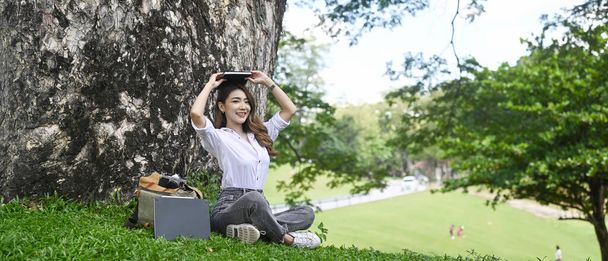 Young asian woman sitting on green grass under tree and smiling to camera. - Φωτογραφία, εικόνα