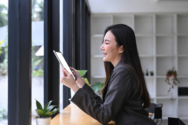 Mujer de negocios sonriente sentada cerca de la ventana en la oficina y usando tableta digital. - Foto, imagen