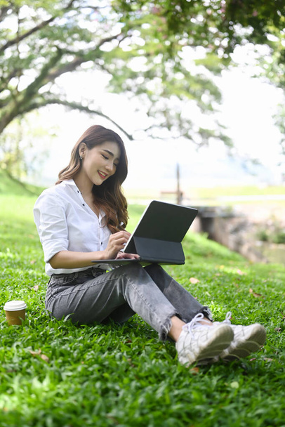 Joven freelancer sentada en el césped en el parque público y trabajando con la tableta de ordenador. - Foto, Imagen