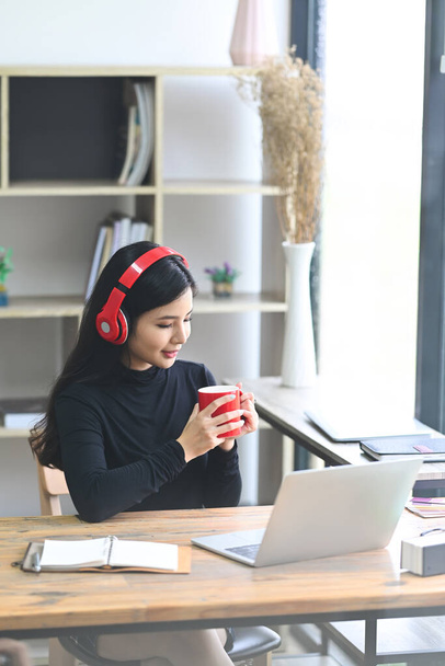 Gülümseyen kadın kulaklıkla müzik dinliyor ve ofiste kahve içiyor.. - Fotoğraf, Görsel