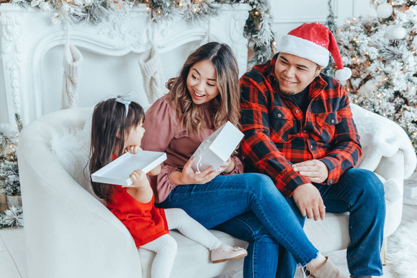 Asyalı aile babası, annesi Noel 'i ya da yeni yılı kutlayan küçük kızına hediye paketi veriyor. Karışık ırk anne, baba, kız evdeki kanepede oturuyor. Kış bayramı kutlaması. - Fotoğraf, Görsel