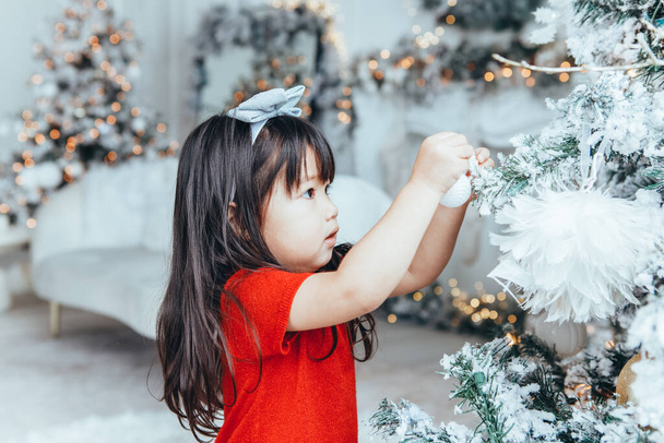 Asian toddler girl child in red dress decorating Christmas tree. Kid celebrating Christmas or New Year at home. Winter holiday celebration. - Фото, изображение
