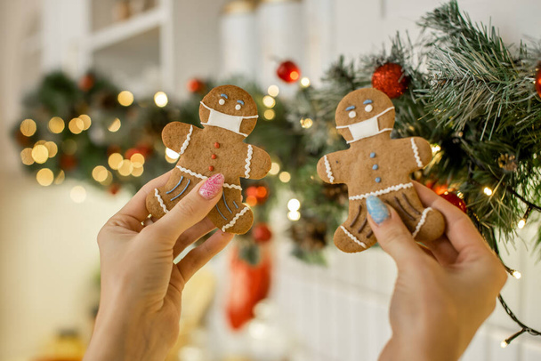 Female hands hold gingerbread man and woman cookies decorated with medical masks on blurred Christmas tree background. Christmas and New Year food concept. - Фото, зображення