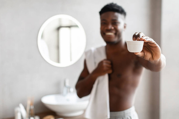 African american man showing moisturizing cream jar in bathroom - Foto, Bild