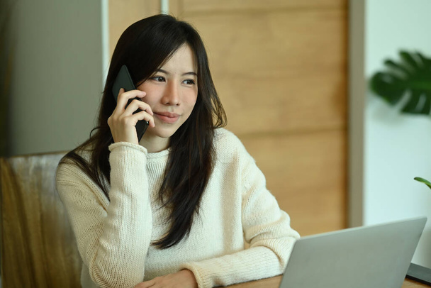 Mladá asijská žena v teplém oblečení pracuje s počítačem notebook a mluví na mobilním telefonu. - Fotografie, Obrázek