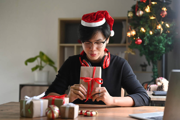 Junger Mann mit Weihnachtsmütze hält Weihnachtsgeschenke mit roter Schleife in der Hand. - Foto, Bild