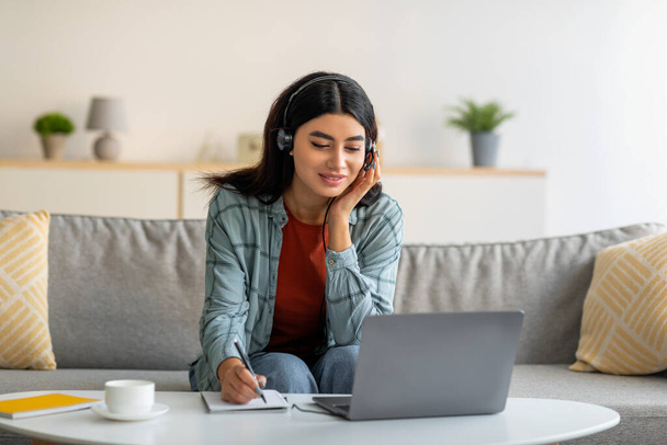 Ev bilgisayarında çalışan, kulaklıklı genç, gülümseyen bir Arap kadın online iş konferansı sırasında bilgi yazıyor. - Fotoğraf, Görsel