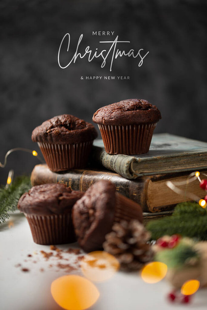 Festive composition with chocolate muffins and ornaments arranged on old books with a Merry Christmas greeting note in the dark background - Foto, Bild
