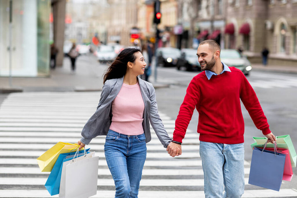 Портрет молодой международной пары с красочными подарочными пакетами, переходящих улицу, держащихся за руки на улице - Фото, изображение